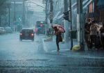 Rua movimentada durante chuva forte, com carros, pedestres e pessoas usando guarda-chuvas.
