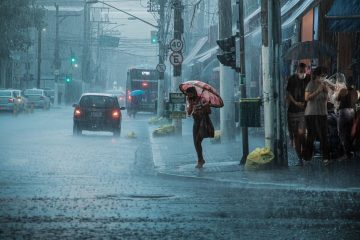 Rua movimentada durante chuva forte, com carros, pedestres e pessoas usando guarda-chuvas.