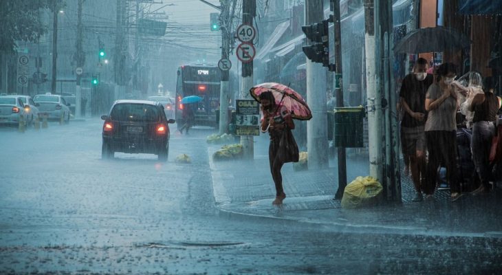 Rua movimentada durante chuva forte, com carros, pedestres e pessoas usando guarda-chuvas.