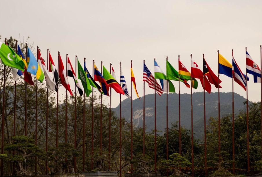 Diversas bandeiras nacionais hasteadas diante de uma paisagem montanhosa, simbolizando a cooperação internacional.