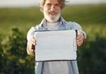 Produtor rural idoso, de barba e óculos, segura uma prancheta e analisa documentos em meio a uma plantação verde.