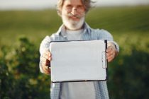 Produtor rural idoso, de barba e óculos, segura uma prancheta e analisa documentos em meio a uma plantação verde.