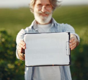 Produtor rural idoso, de barba e óculos, segura uma prancheta e analisa documentos em meio a uma plantação verde.