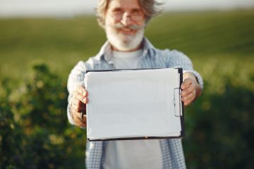 Produtor rural idoso, de barba e óculos, segura uma prancheta e analisa documentos em meio a uma plantação verde.