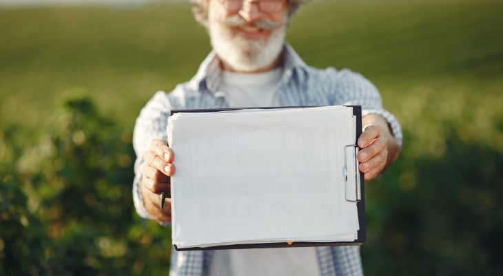Produtor rural idoso, de barba e óculos, segura uma prancheta e analisa documentos em meio a uma plantação verde.